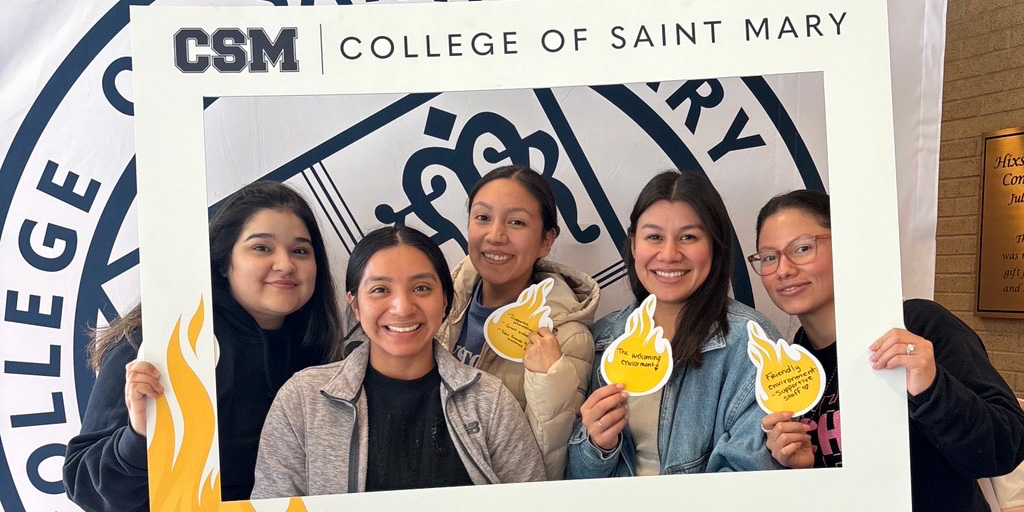 Five College of Saint Mary students pose with a CSM frame in front of the CSM seal.